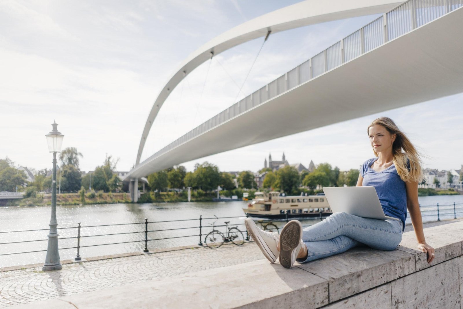 Netherlands, Maastricht, young woman sitting on a wall at the riverside with laptop